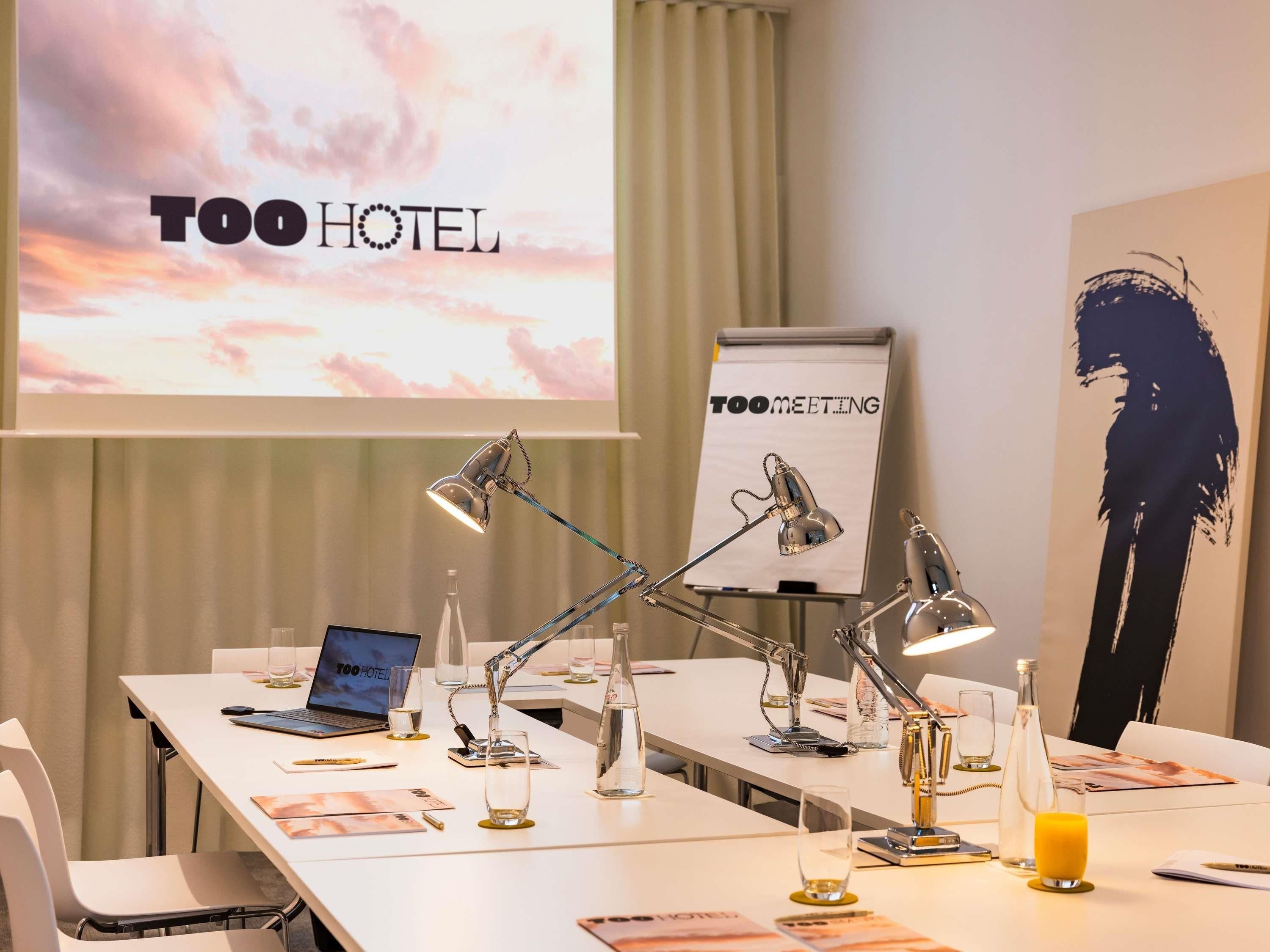 A conference room with a laptop, multiple desk lamps, a projector screen displaying 'TOO HOTEL', and a sign that reads 'TOO MEETING'.