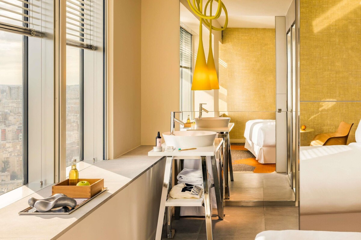 A modern hotel room with a large window, a desk with a sink, and a bed.