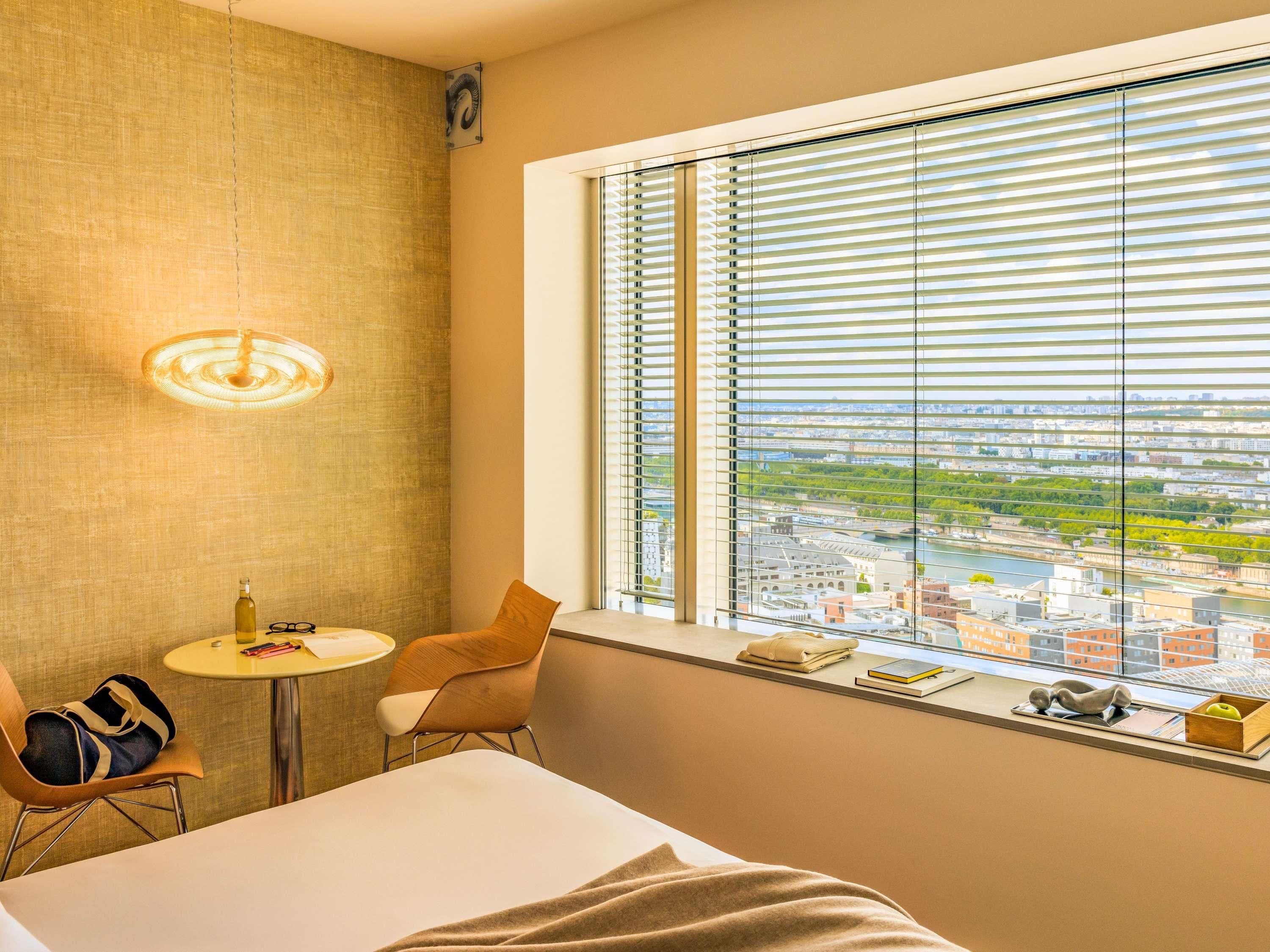 Habitación de hotel con cama, una mesita, una silla, ventana con persianas y vista a la ciudad.