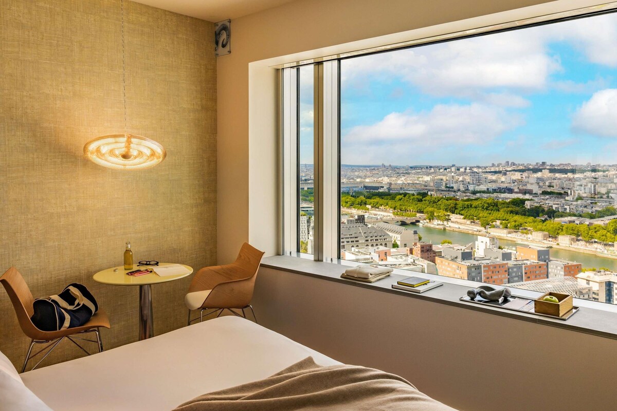 A hotel room with a large window, a bed, a small table, and a chair.