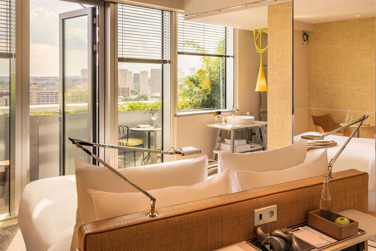 A hotel room with a bed, a desk with a lamp, a small table with a chair, a window with blinds, and a view of buildings and greenery.