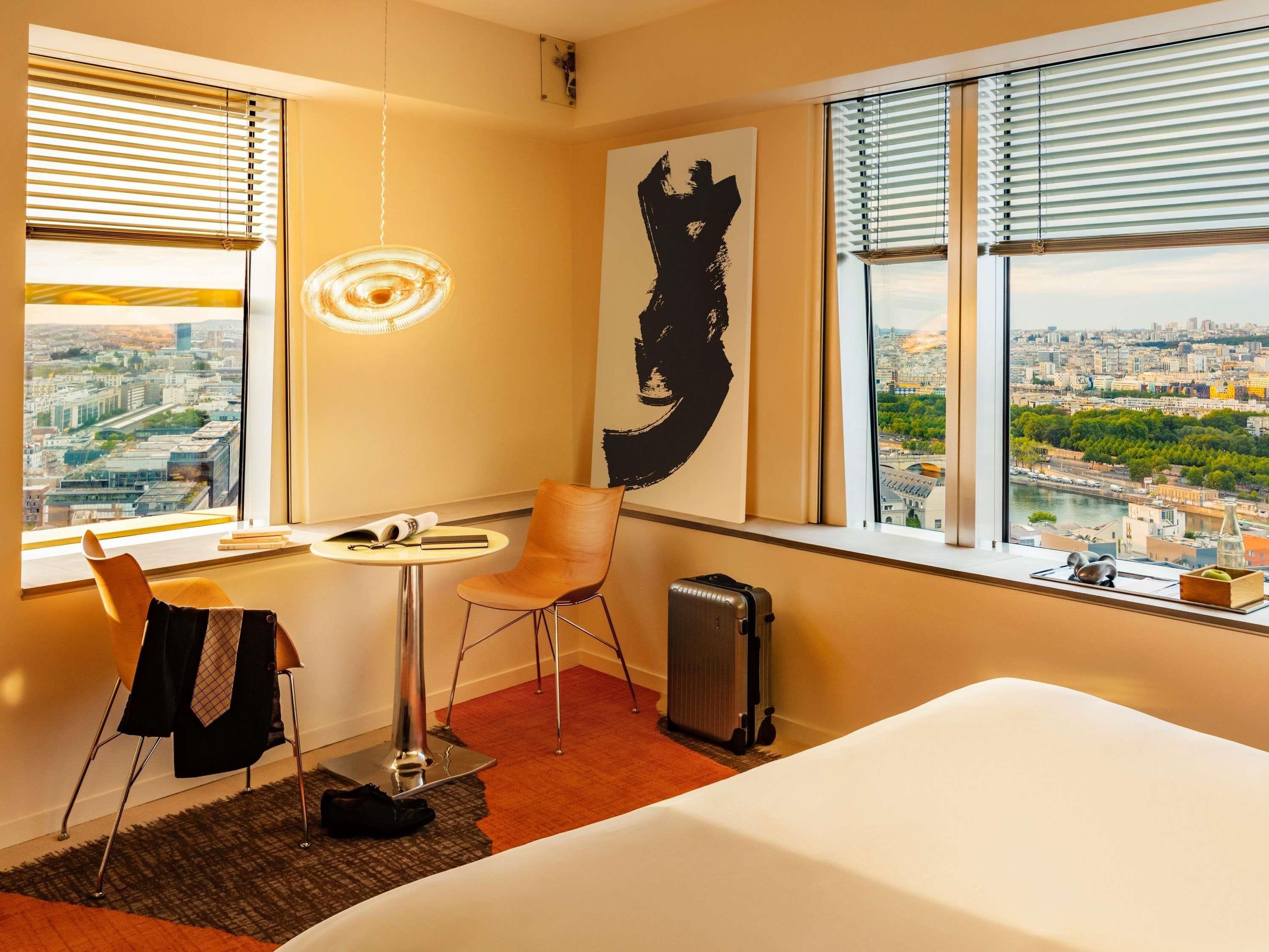 Ein Hotelzimmer mit Bett, Stuhl, Schreibtisch, Koffer und einem großen Fenster mit Blick auf die Stadt.