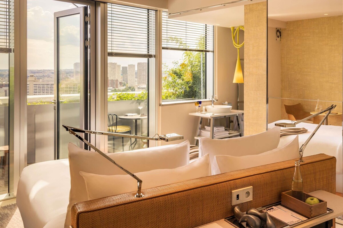 A hotel room with a bed, a desk with a lamp, a small table with a chair, a window with blinds, and a view of buildings and greenery.