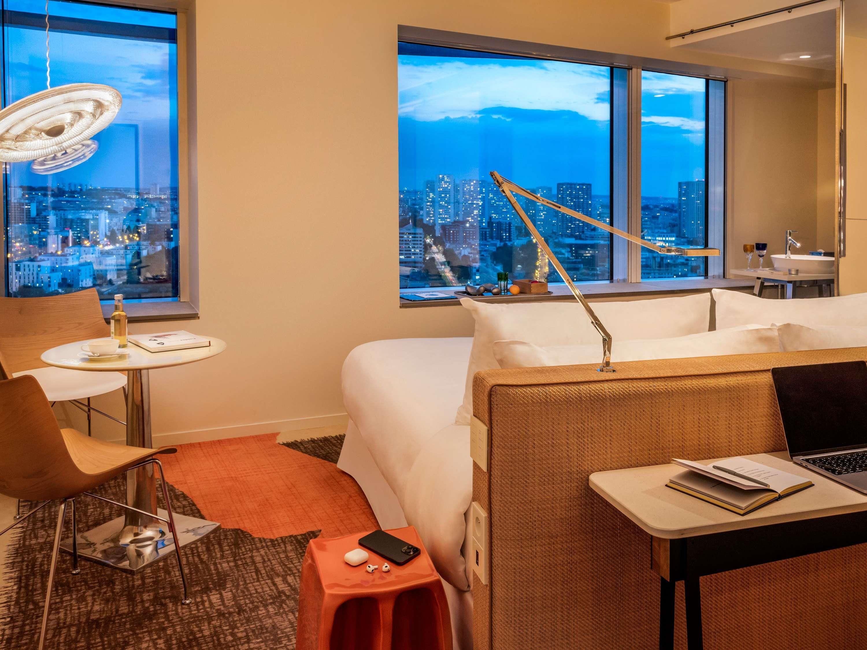 A hotel room with a city view, a desk with a laptop, a chair, a small table with a bottle, and a bedside table with a book and a phone.