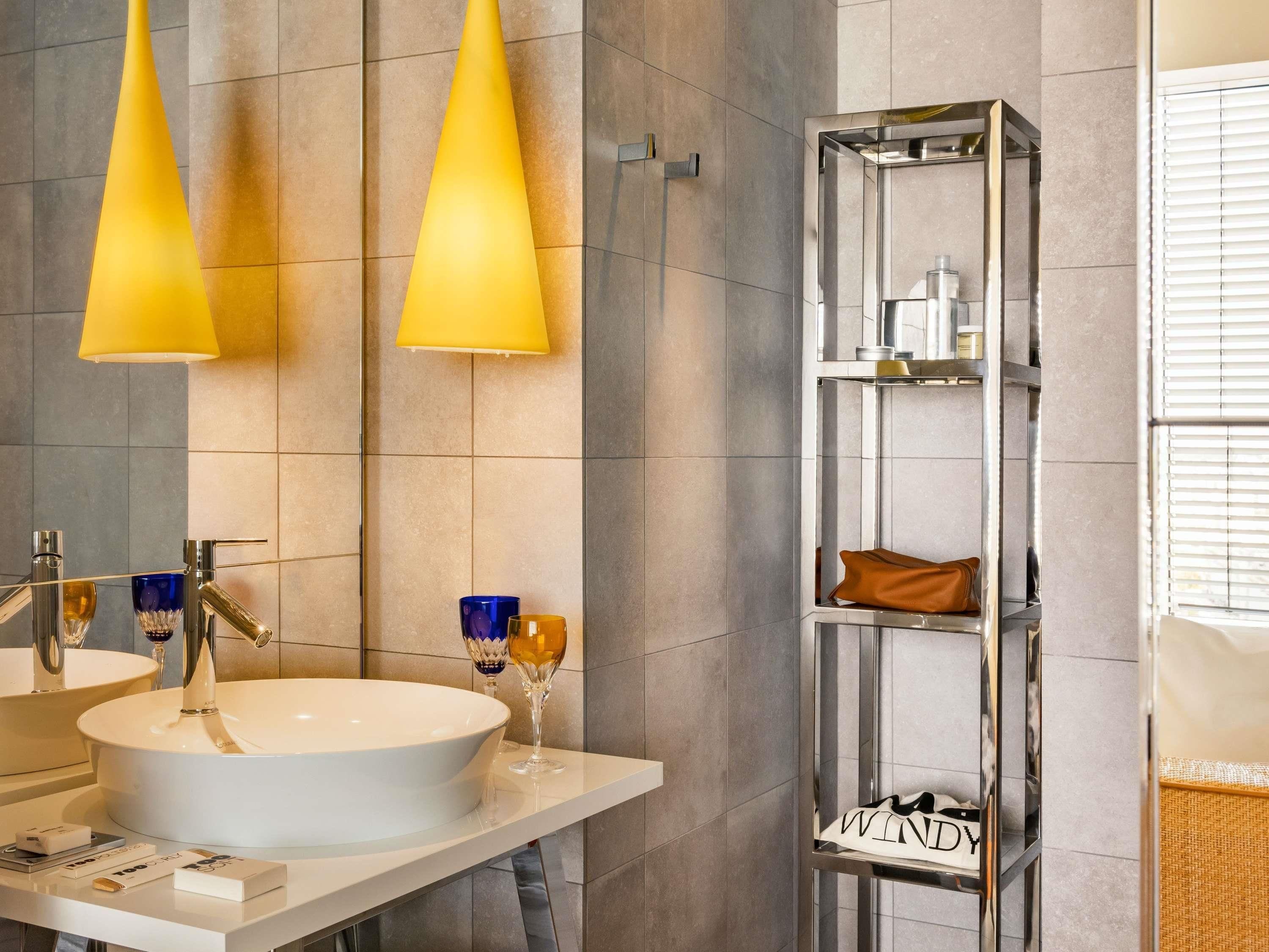 A modern bathroom with a white sink, two yellow pendant lights, a mirror, and a tall, narrow shelf unit with a leather item and a shoe.