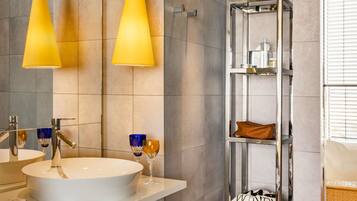 A modern bathroom with a white sink, two yellow pendant lights, a mirror, and a tall, narrow shelf unit with a leather item and a shoe.