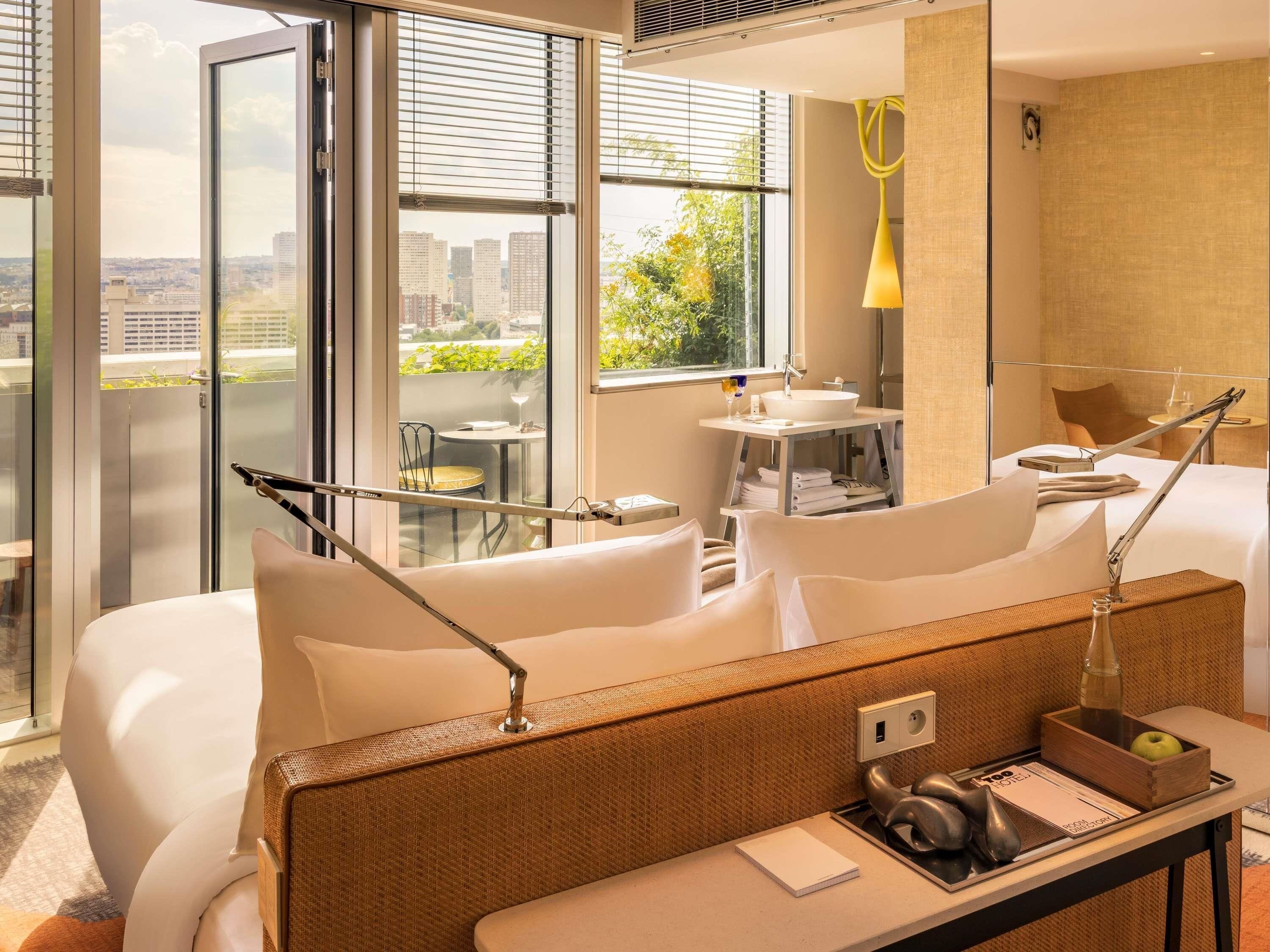A modern hotel room with a large window, a private balcony, a bed, a desk with a lamp, and a small table with a vase.