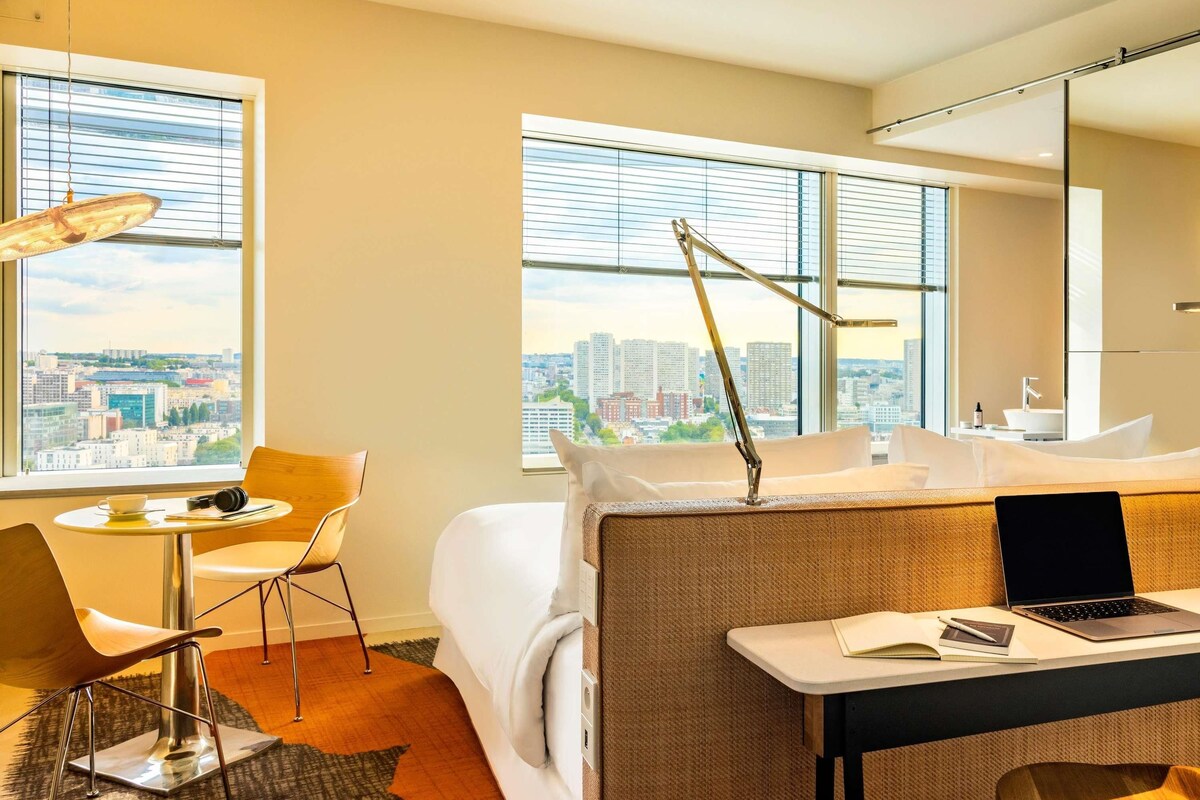 A modern hotel room with a city view, a bed, a desk with a laptop, a chair, and a lamp.