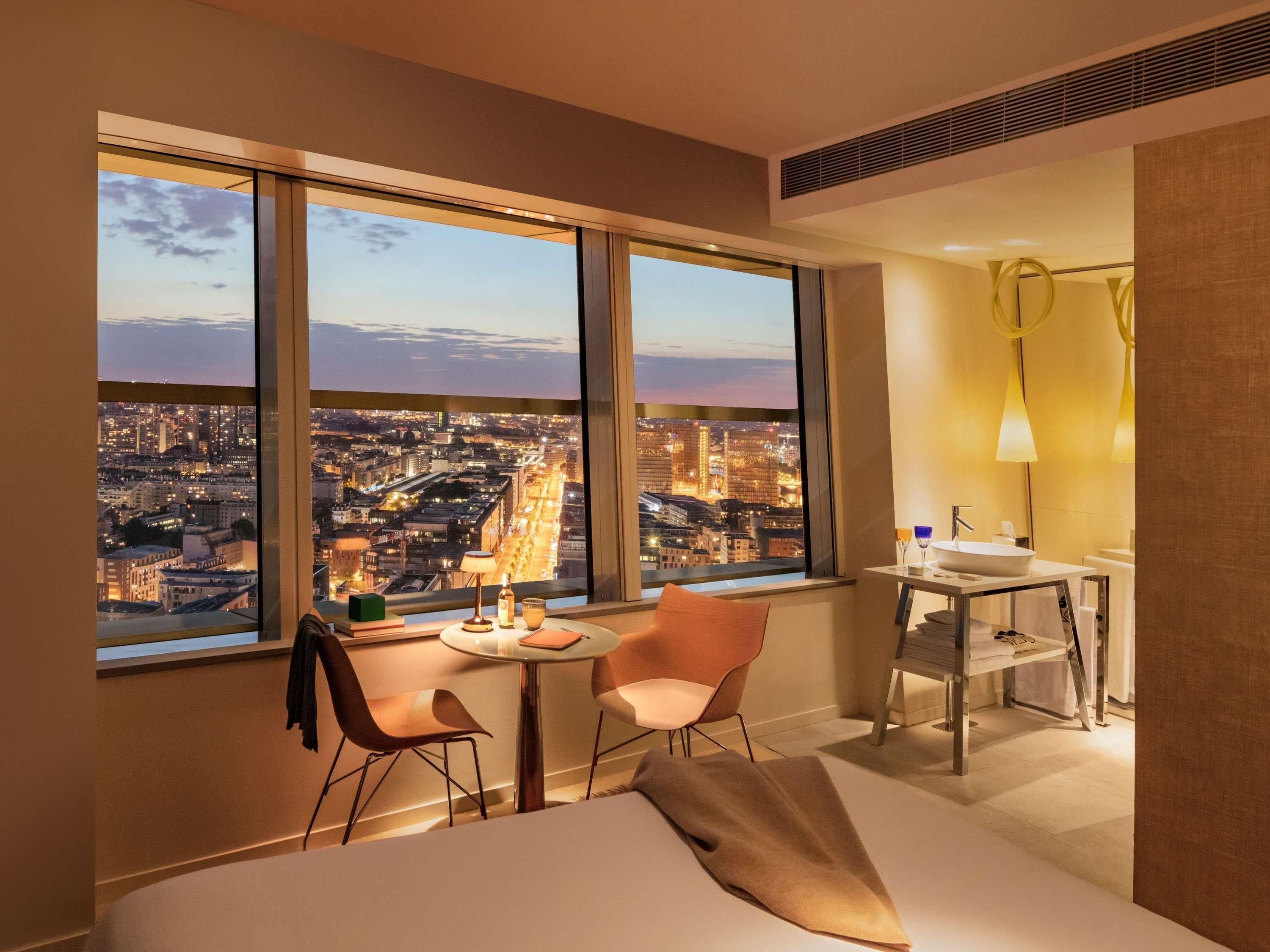 Ett modernt hotellrum med utsikt över staden, en säng, ett litet bord med en lampa och ett skrivbord med en handfat.