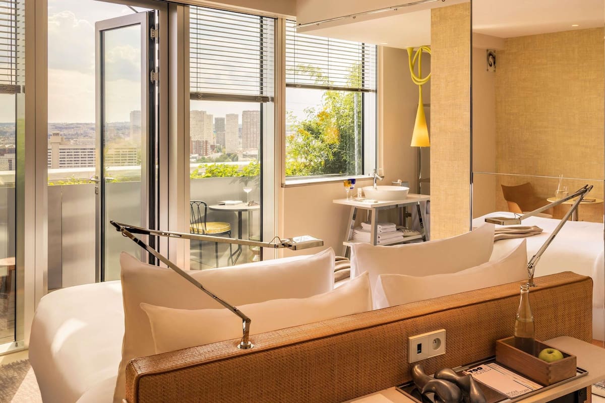 A hotel room with a bed, a desk with a lamp, a small table with a chair, a window with blinds, and a view of buildings and greenery.