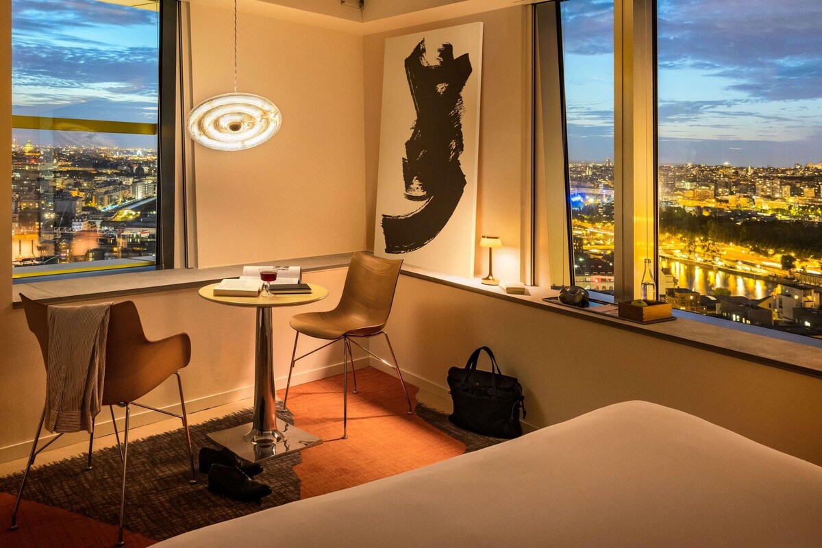 A hotel room with a city view, a round table, a chair, a bed, and a large abstract painting.