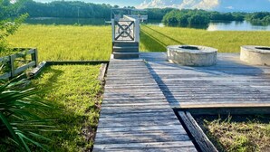 Un muelle de madera que lleva a un pequeño espigón con una puerta metálica, rodeado de exuberantes campos verdes y un lago.