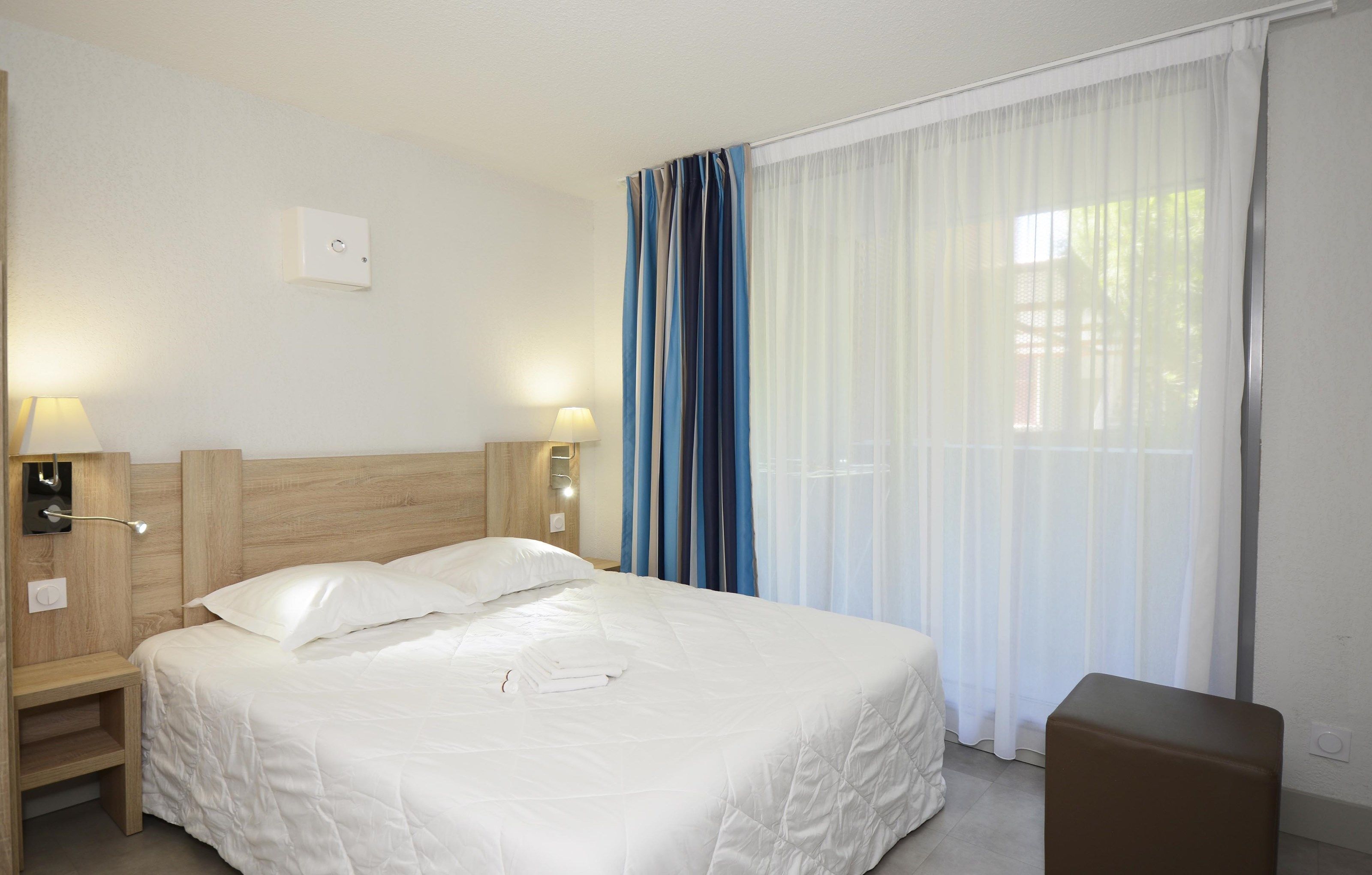 A hotel room with a wooden headboard, two bedside lamps, a bed with white linens, a brown ottoman, and sheer curtains.