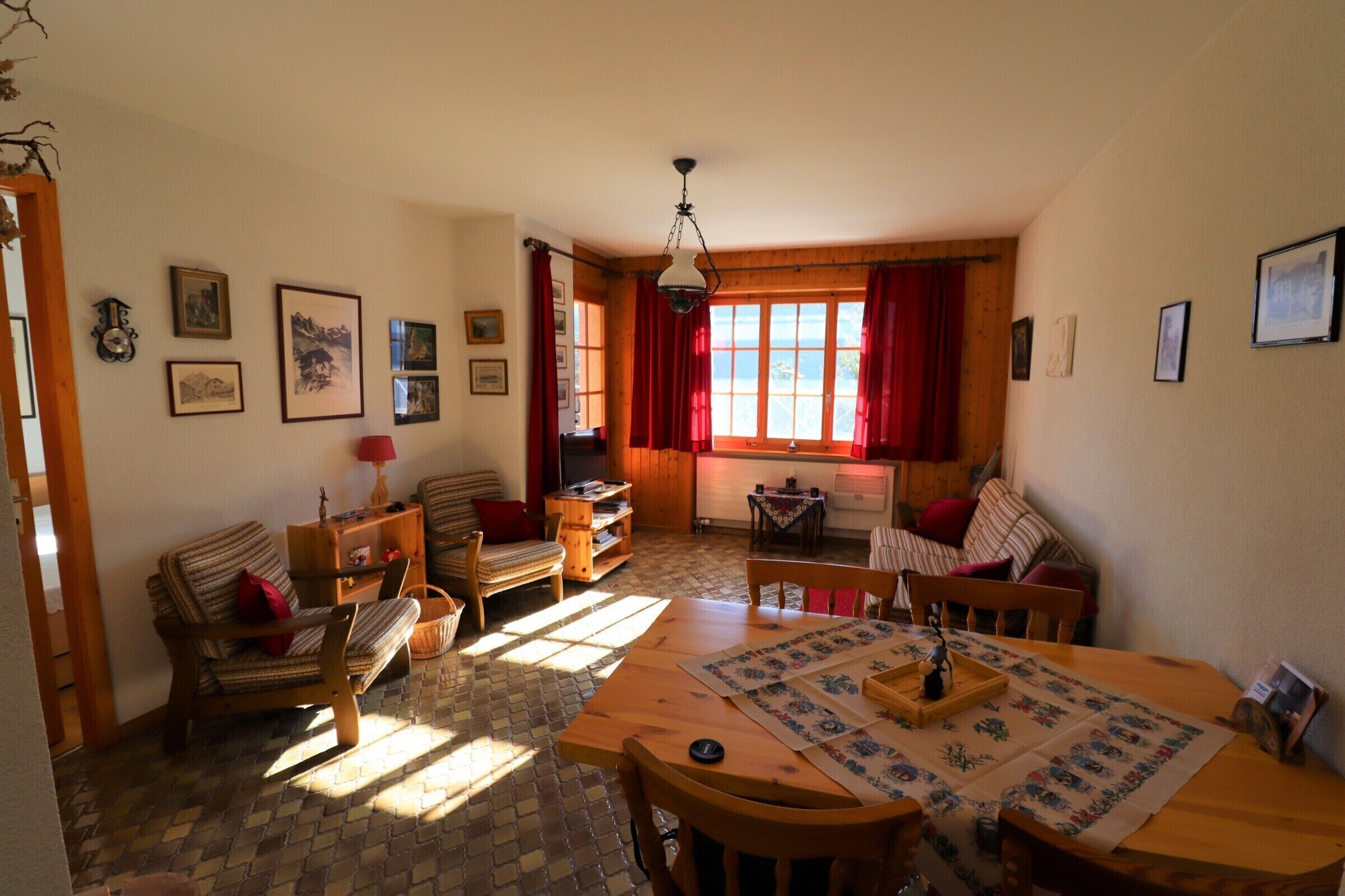 Un salon chaleureux avec une table en bois, des chaises et un canapé. Les murs sont ornés de tableaux encadrés et une fenêtre est agrémentée de rideaux rouges.