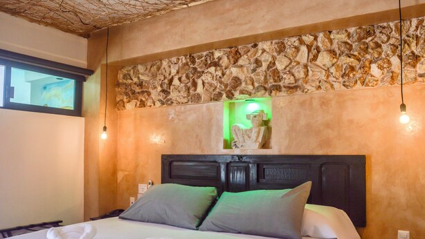 A hotel room with a stone wall, a bed with green pillows, and a nightstand with a lamp. - Habitaciones Colonial Zaci (Valladolid)
