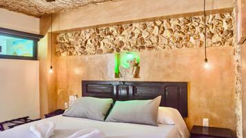 A hotel room with a stone wall, a bed with two pillows, a nightstand, and a wall-mounted light fixture.