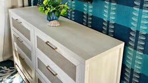 Een moderne woonkamer met een flatscreen televisie aan een wand, een houten dressoir met lades, een potplant en behang met een patroon.
