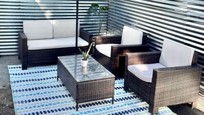 Een buitenterras met rieten meubilair, een salontafel met glazen blad en een blauwe parasol.