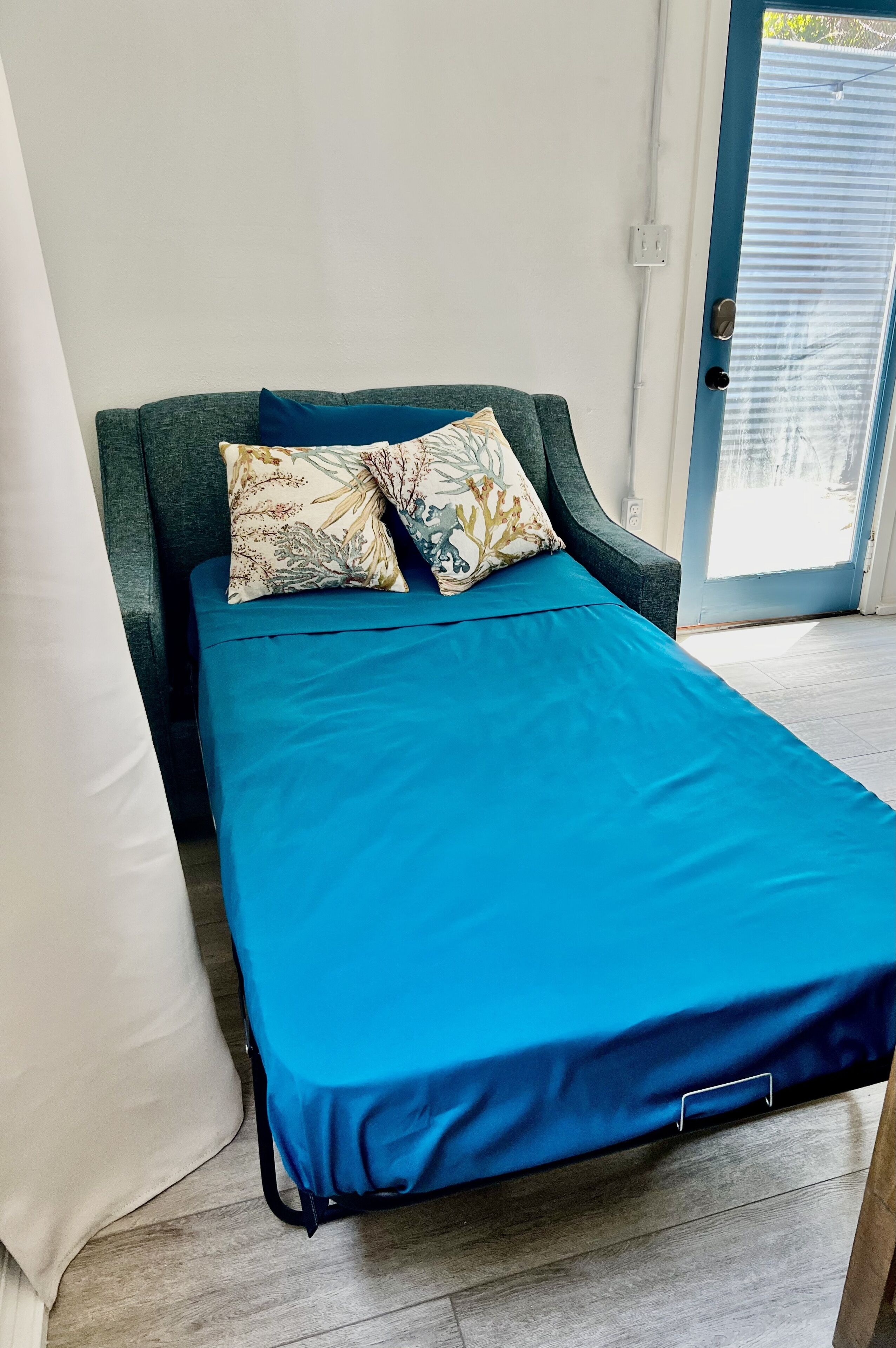 Een bed met een blauwe matras, bloemenkussens, een wit kussen en een donkergroen hoofdeinde.