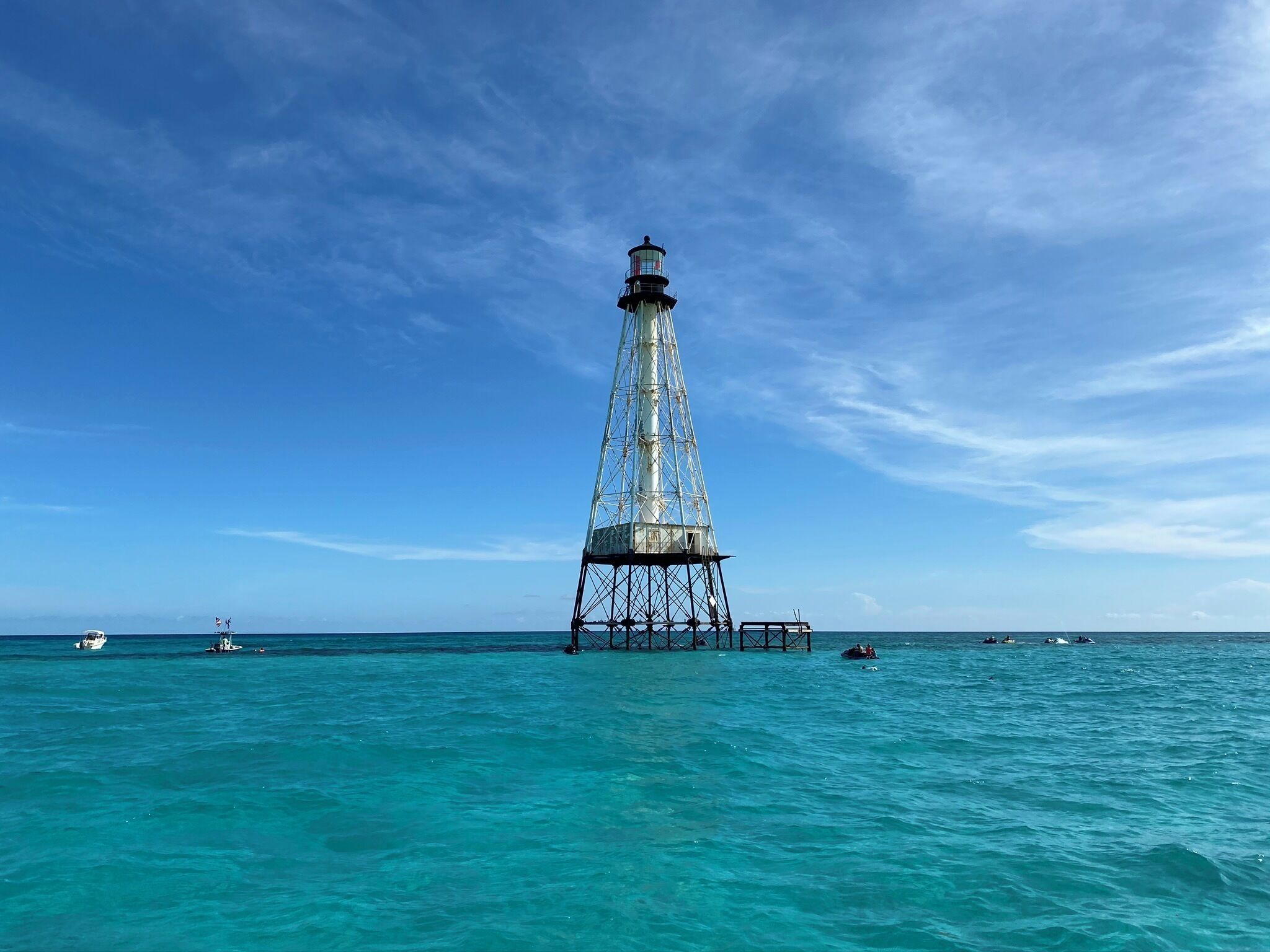 Ein Leuchtturm mit Metallgerüst und einem Glaslaternenraum, umgeben von ruhigem türkisblauem Wasser und blauem Himmel.