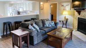 A living room with a sofa, coffee table, and stools. There is a kitchen area with cabinets and a sink. A fireplace is visible in the background. - Family Friendly Pines Condo-3b/2b (Pinetop-Lakeside)