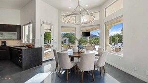 Una cocina moderna con armarios oscuros, una isla grande y un comedor con lámpara colgante y ventanas amplias con vistas al exterior.