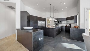 Una cocina moderna con armarios oscuros, una isla central con un lavabo, iluminación colgante y vistas al área de estar contigua.