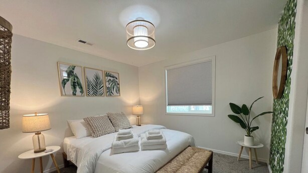 A bedroom with a bed, two bedside tables, a bench, a hanging light fixture, a window with blinds, and a potted plant. - Enjoy a stylish experience at The Franklin (Pasco)