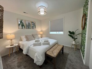 A bedroom with a bed, two bedside tables, a bench, a hanging light fixture, a window with blinds, and a potted plant. - Enjoy a stylish experience at The Franklin (Pasco)