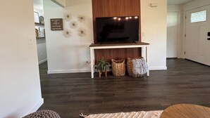 A living room with a flat-screen TV on a white stand, dark wooden flooring, and a white door in the background. - Enjoy a stylish experience at The Franklin (Pasco)