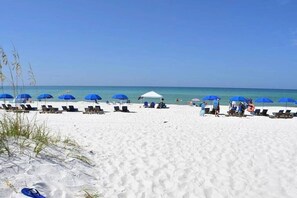 A beach with rows of sun loungers and blue umbrellas, a white tent, and clear blue skies.