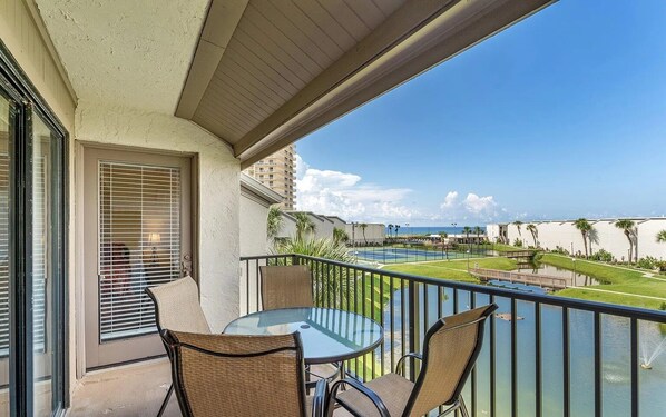 A balcony with a glass table and chairs, overlooking a pool and a view of the ocean.