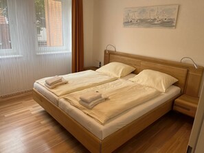 Uma cama de casal com duas almofadas, uma mesa de cabeceira, uma janela com cortinas e um quadro na parede.