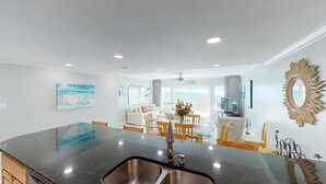 A modern kitchen with a large island, stainless steel sinks, and a dining area with a glass table and chairs. - Island Time Gulf-Front Townhome/ Pet Friendly (Navarre Beach)