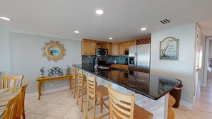 A modern kitchen with wooden cabinets, a black countertop, and stainless steel appliances. There is a dining area with wooden chairs and a table. - Island Time Gulf-Front Townhome/ Pet Friendly (Navarre Beach)