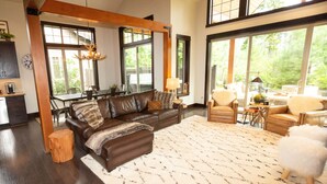 A spacious living room with a large brown leather sofa, a white shag rug, and a wooden coffee table. There are two windows providing natural light and a view of greenery outside. - FAll in Suncadia! Walk to the new Suncadia social & Golf! Hot Tub and Games! (Cle Elum)