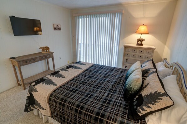 Een slaapkamer met een bed, nachtkastje, lamp, kast, televisie aan de muur en een raam met jaloezieën.