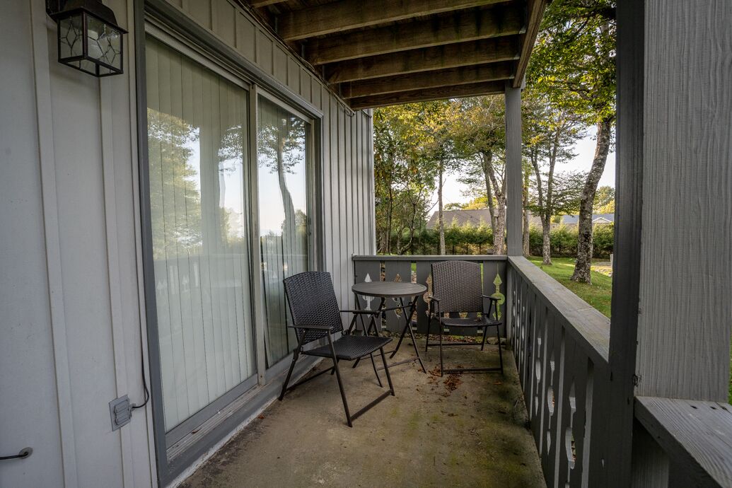 A covered outdoor patio with a table and chairs, a lantern, and a view of trees and a house in the background.