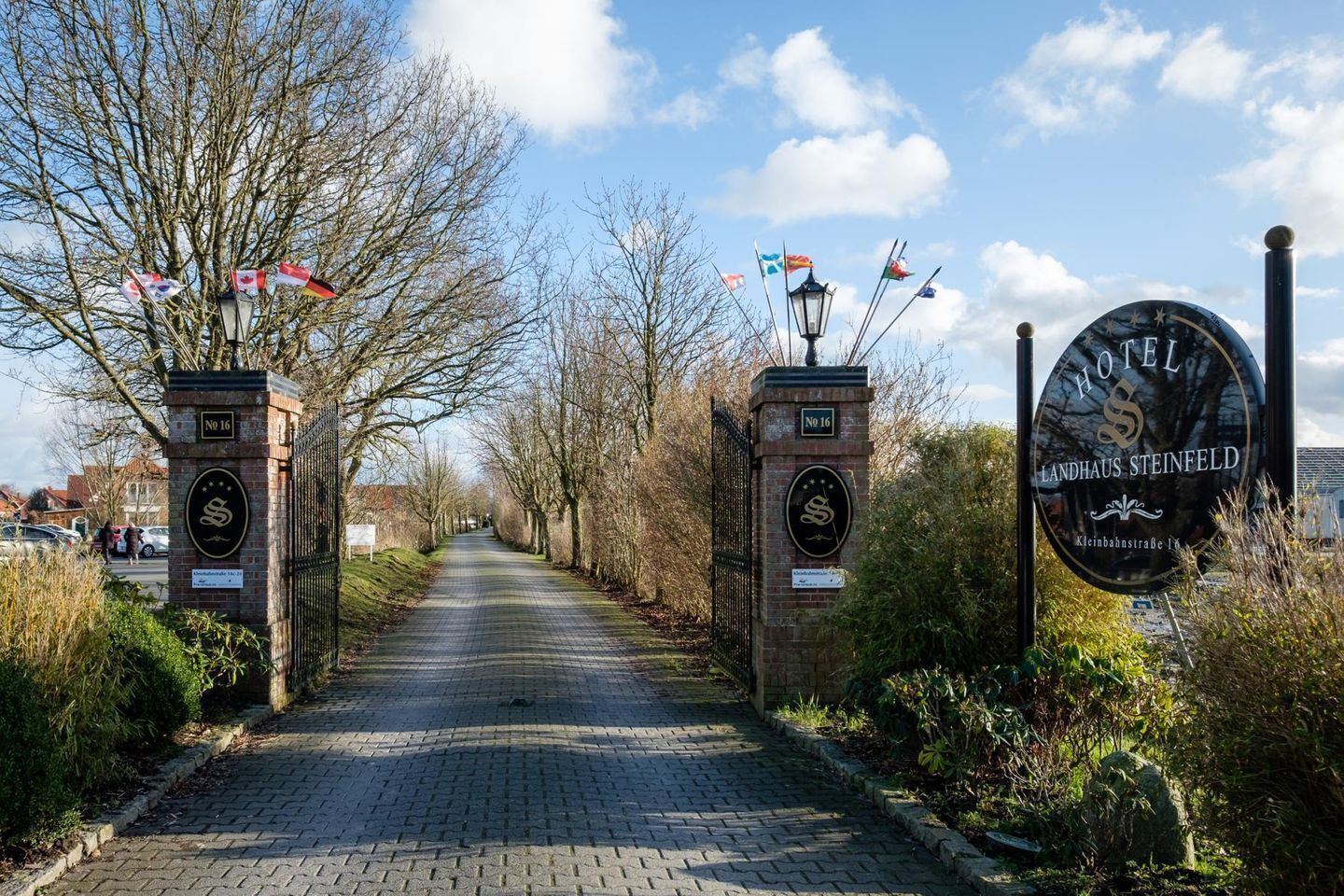 Ein Hotel Eingang mit einem gepflasterten Weg, zwei Ziegelstützsäulen mit Fahnen und einem runden Schild mit dem Namen des Hotels.