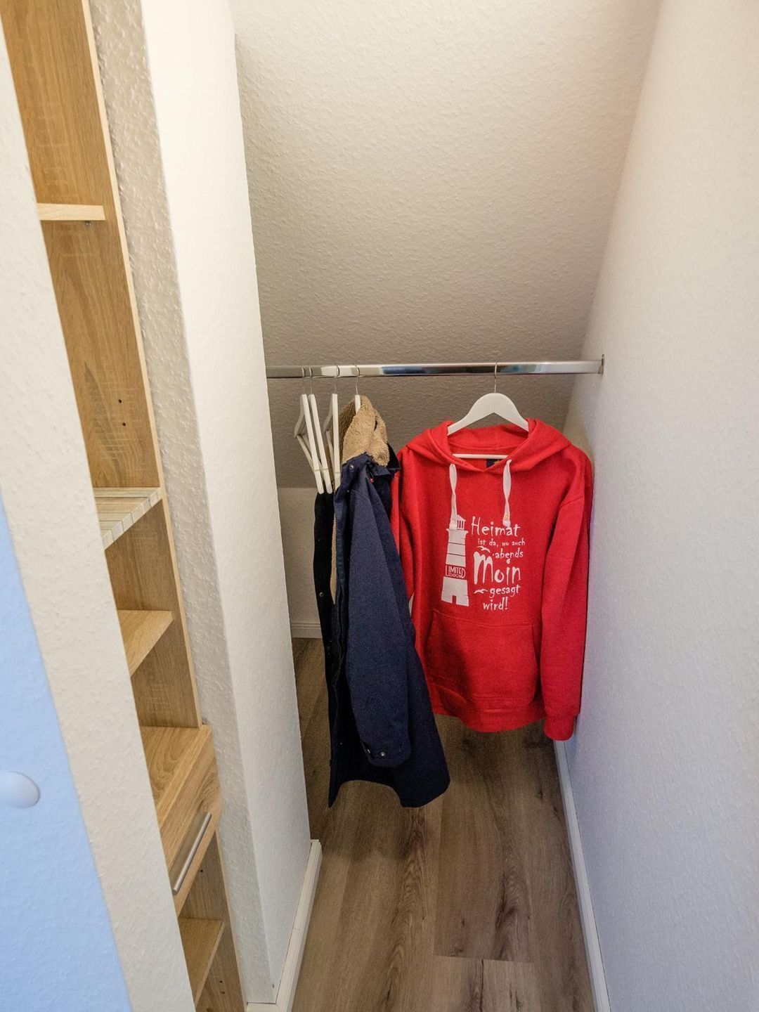 A narrow hallway with a wooden staircase on the left, a metal handrail, and a clothing rack with a red hoodie and a blue jacket.