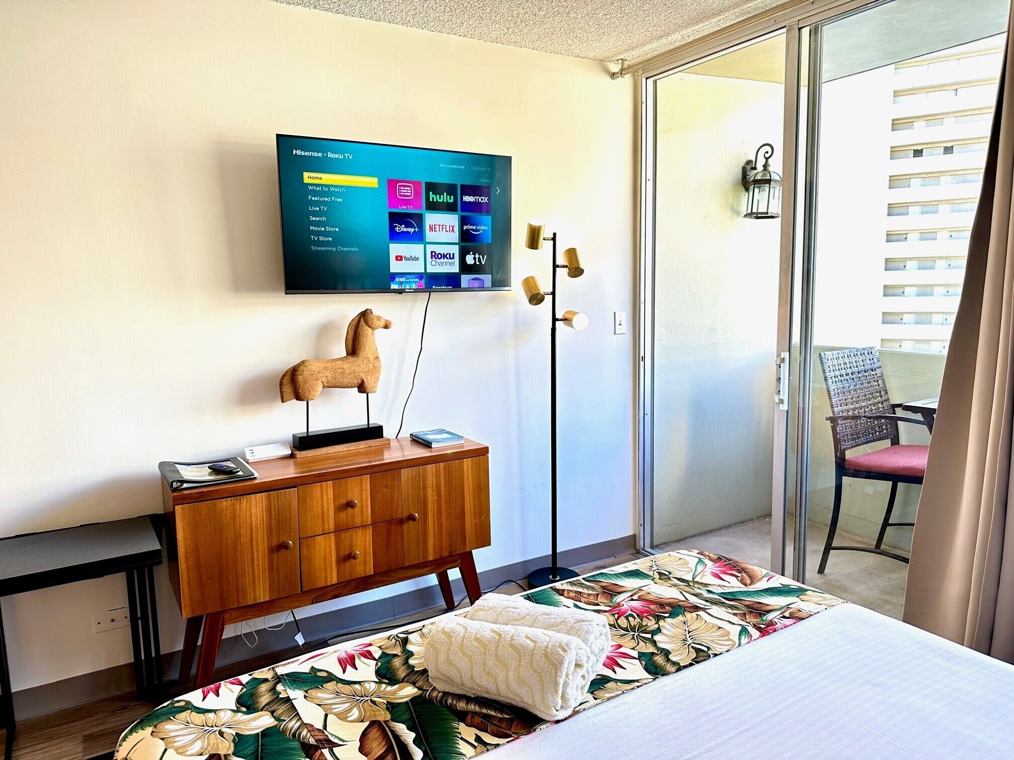 A hotel room with a bed, a wooden dresser, a mounted TV displaying streaming services, a decorative horse sculpture, and a view of a building through a glass door.