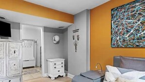 Een slaapkamer met een bed, een witte dressoir, een nachtkastje, een wandklok en een kleurrijk abstract schilderij.