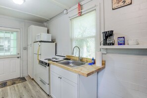 Eine kompakte Küche mit weißen Schränken, einem Herd, einer Mikrowelle und einem Spülbecken. Es gibt ein Fenster mit Jalousien, ein Regal mit einer Kaffeemaschine und einer Dose Maxwell House Kaffee.