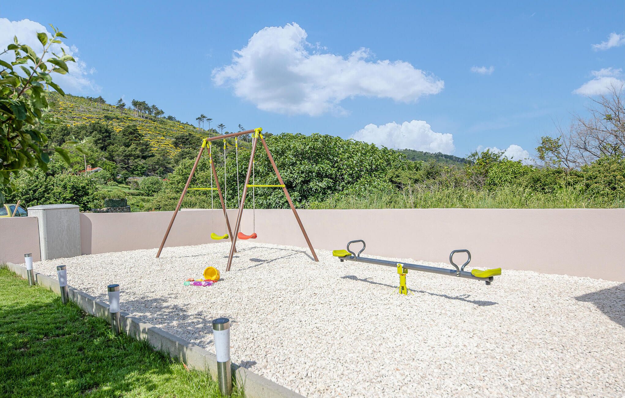 A playground with a swing set and seesaw on a gravel surface, surrounded by greenery and hills.