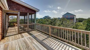 A wooden deck with a railing, a covered area with a chair, and a view of other buildings in the background. - New construction home short walk to the beach! (Avon)