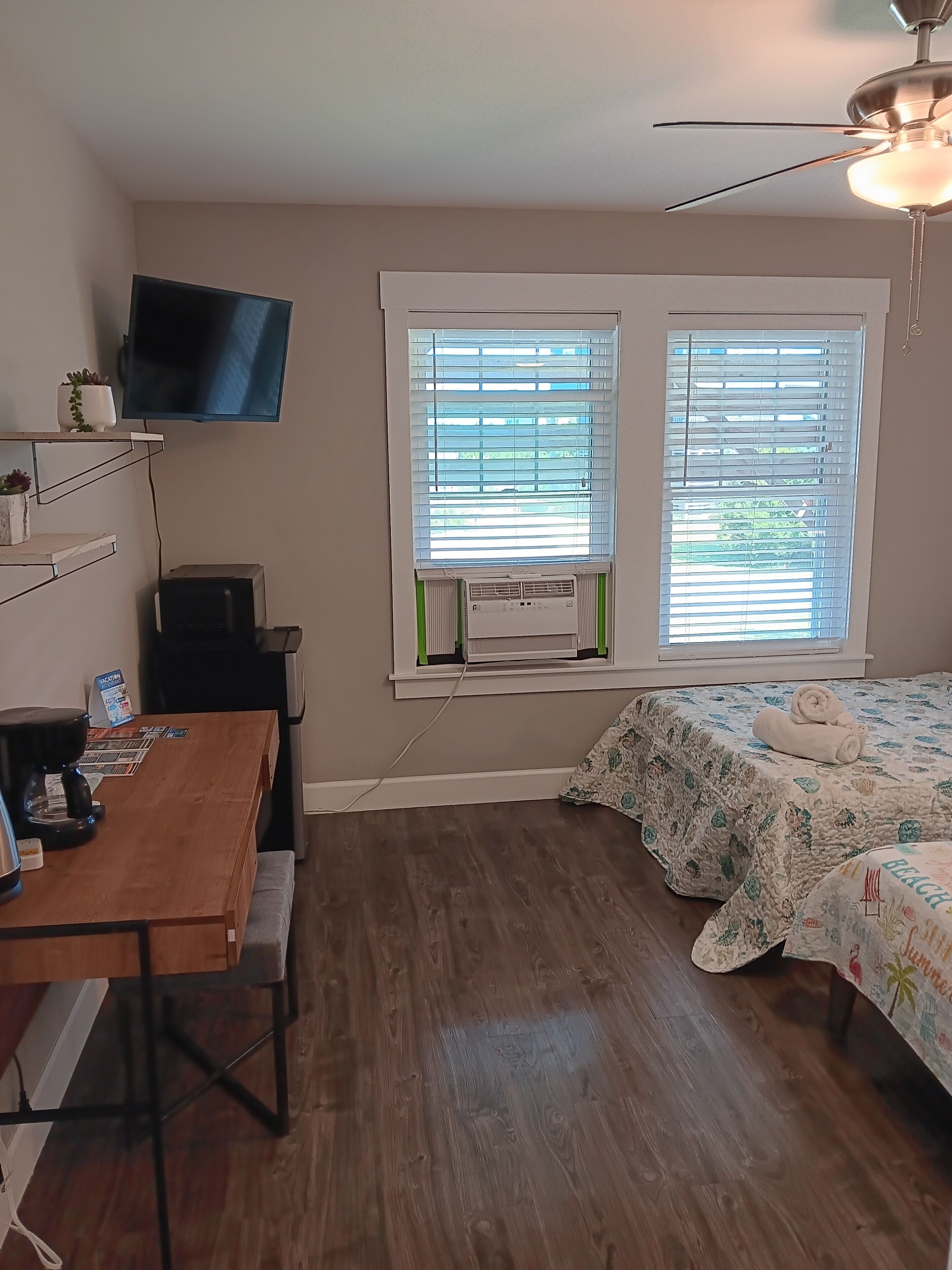 A bedroom with a wooden floor, a bed with a floral bedspread, a desk with a coffee maker, a television mounted on the wall, and a window with blinds.