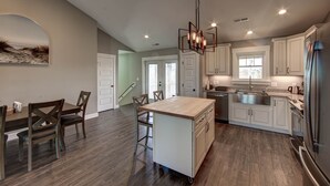 A modern kitchen with white cabinetry, a central island, stainless steel appliances, and a dining area with wooden chairs. - New construction home short walk to the beach! (Avon)