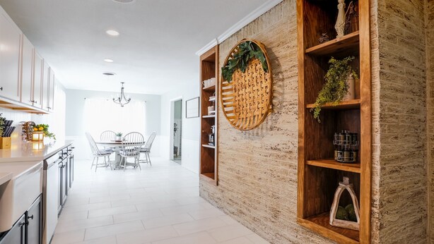 A modern kitchen with white cabinetry, a dining area with a chandelier, and a brick accent wall with shelves. - Melanie Park Raider Cottagecore Design 4/3 (Lubbock)