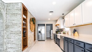 A modern kitchen with white cabinets, a brick accent wall, and a wooden shelf unit. - Melanie Park Raider Cottagecore Design 4/3 (Lubbock)
