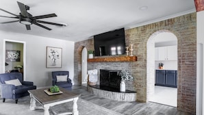 A living room with a fireplace, two armchairs, a coffee table, and a TV mounted above the fireplace. - Melanie Park Raider Cottagecore Design 4/3 (Lubbock)